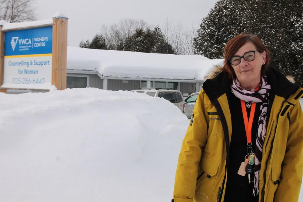 Paula Pepping wearing a bright yellow coat, standing next to YWCA Haliburton County's street sign on a snowy day