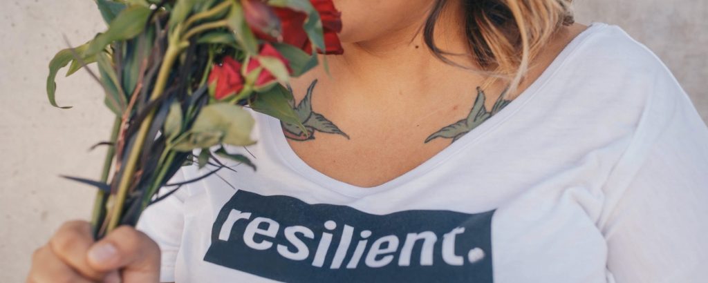 a tatooed woman smelling flowers, wearing a white shirt that reads "resilient"