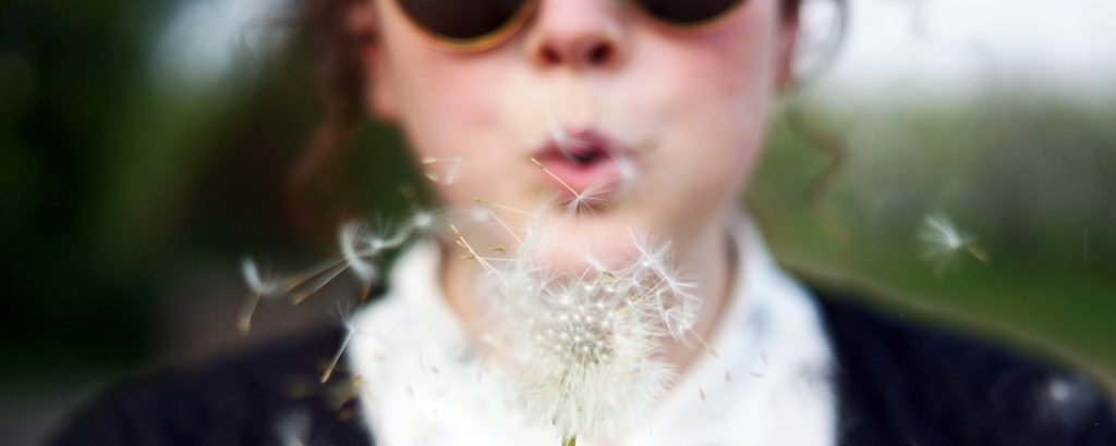a woman wearing sunglasses, making a wish on a fluffy white dandelion