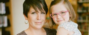 a woman with short hair and hoop earrings holds a young girl wearing pink glasses on her hip
